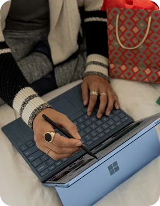 Een close-up van iemand die met een stylus-pen op het scherm van een blauwe Microsoft Surface typt en navigeert, met een papieren cadeautas op de achtergrond